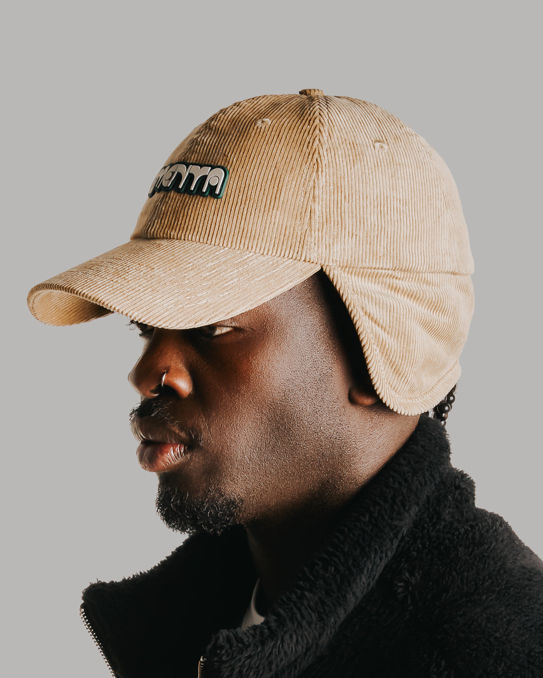 A six-panel cap constructed with a corduroy outer shell. The design includes a continuous flap extending from ear to ear with a sherpa interior for insulation and comfort, covering the back of the neck for full cold-weather protection. The flaps can be worn up or down, allowing for adjustable coverage. The cap features a curved brim for structure and coverage. The front panel displays the Dots Ementa logo.