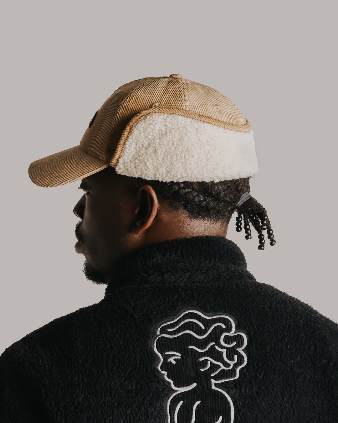 A six-panel cap constructed with a corduroy outer shell. The design includes a continuous flap extending from ear to ear with a sherpa interior for insulation and comfort, covering the back of the neck for full cold-weather protection. The flaps can be worn up or down, allowing for adjustable coverage. The cap features a curved brim for structure and coverage. The front panel displays the Dots Ementa logo.