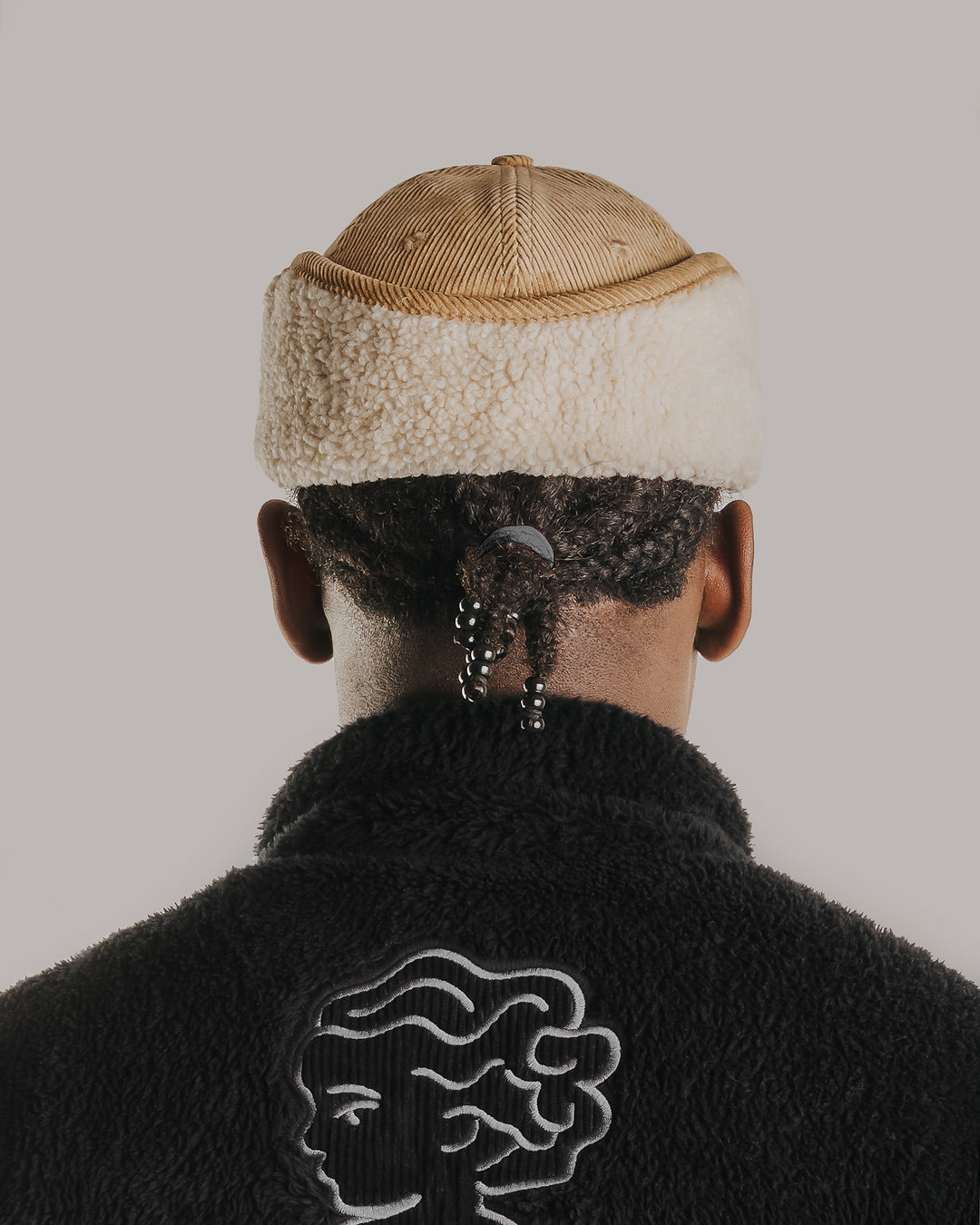 A six-panel cap constructed with a corduroy outer shell. The design includes a continuous flap extending from ear to ear with a sherpa interior for insulation and comfort, covering the back of the neck for full cold-weather protection. The flaps can be worn up or down, allowing for adjustable coverage. The cap features a curved brim for structure and coverage. The front panel displays the Dots Ementa logo.