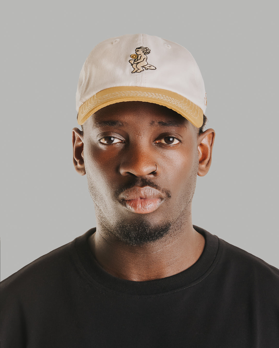 Off-white crown, camel brim, white stitching holding it all together. The Ikon Flower is embroidered on the front, the kind of detail that makes you look twice. On the side, "Moved by Friendship" is stitched in, a reminder of what really matters. Around back, the Dots logo sits above the adjustable strap—small details that tie it all together.
