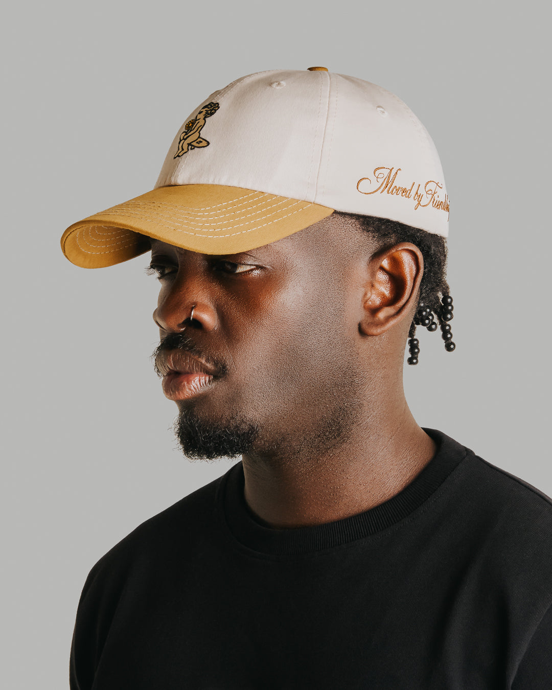 Off-white crown, camel brim, white stitching holding it all together. The Ikon Flower is embroidered on the front, the kind of detail that makes you look twice. On the side, "Moved by Friendship" is stitched in, a reminder of what really matters. Around back, the Dots logo sits above the adjustable strap—small details that tie it all together.