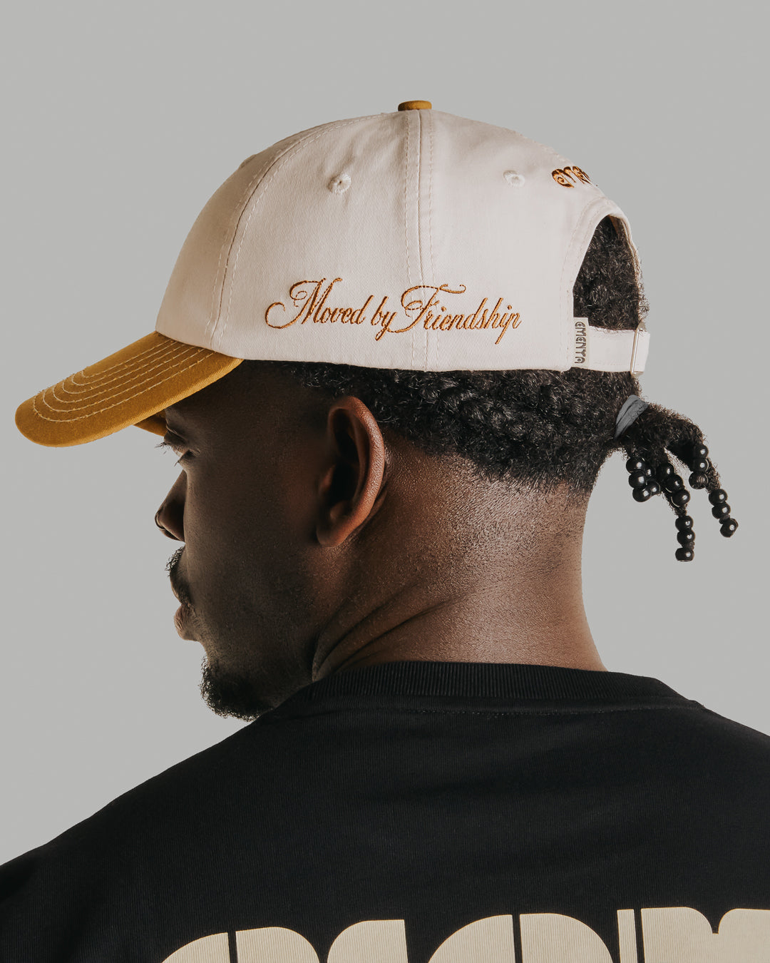 Off-white crown, camel brim, white stitching holding it all together. The Ikon Flower is embroidered on the front, the kind of detail that makes you look twice. On the side, "Moved by Friendship" is stitched in, a reminder of what really matters. Around back, the Dots logo sits above the adjustable strap—small details that tie it all together.