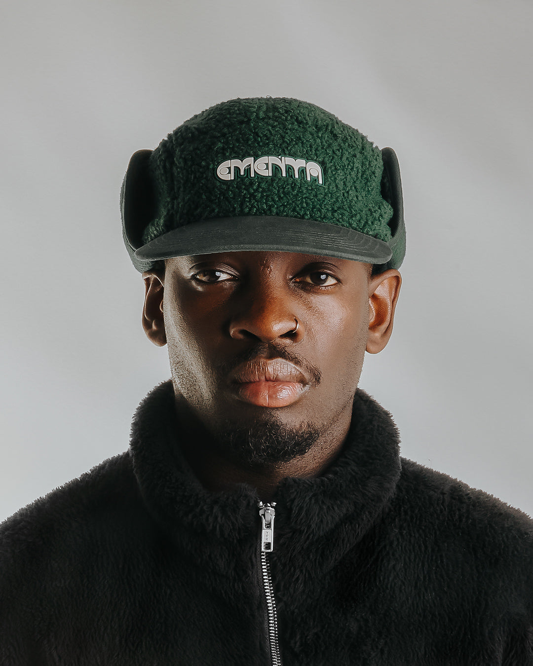 The Oreo Cap is a six-panel cap constructed with a sherpa outer shell for insulation and texture. It features ear flaps with a polar fleece lining on the inside for additional warmth and comfort. The flaps can be worn up or down, allowing for adjustable coverage. The cap has a curved brim that maintains its shape. The front panel displays the Dots Ementa logo.