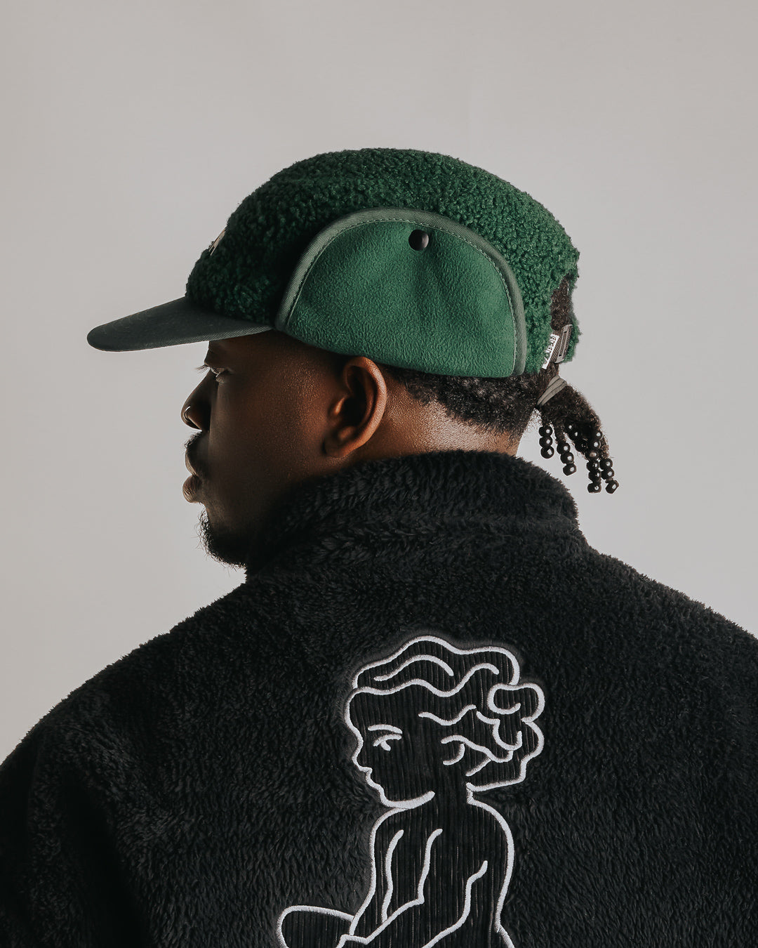 The Oreo Cap is a six-panel cap constructed with a sherpa outer shell for insulation and texture. It features ear flaps with a polar fleece lining on the inside for additional warmth and comfort. The flaps can be worn up or down, allowing for adjustable coverage. The cap has a curved brim that maintains its shape. The front panel displays the Dots Ementa logo.