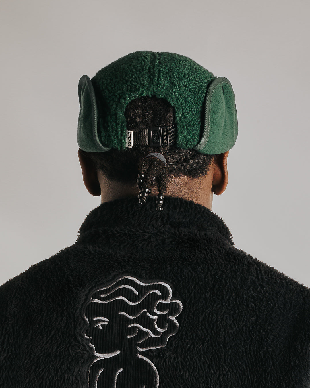 The Oreo Cap is a six-panel cap constructed with a sherpa outer shell for insulation and texture. It features ear flaps with a polar fleece lining on the inside for additional warmth and comfort. The flaps can be worn up or down, allowing for adjustable coverage. The cap has a curved brim that maintains its shape. The front panel displays the Dots Ementa logo.