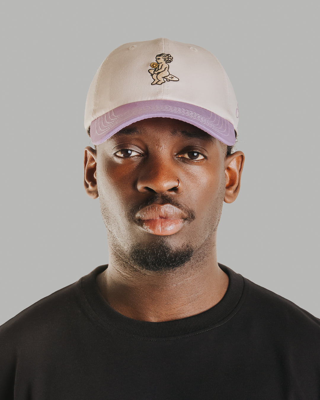 Off-white crown, lilac brim, white stitching holding it all together. The Ikon Flower is embroidered on the front, the kind of detail that makes you look twice. On the side, "Moved by Friendship" is stitched in, a reminder of what really matters. Around back, the Dots logo sits above the adjustable strap—small details that tie it all together.