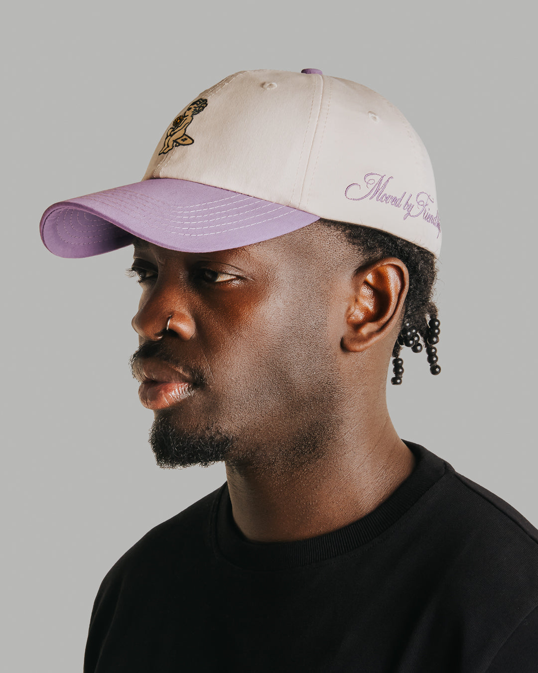 Off-white crown, lilac brim, white stitching holding it all together. The Ikon Flower is embroidered on the front, the kind of detail that makes you look twice. On the side, "Moved by Friendship" is stitched in, a reminder of what really matters. Around back, the Dots logo sits above the adjustable strap—small details that tie it all together.