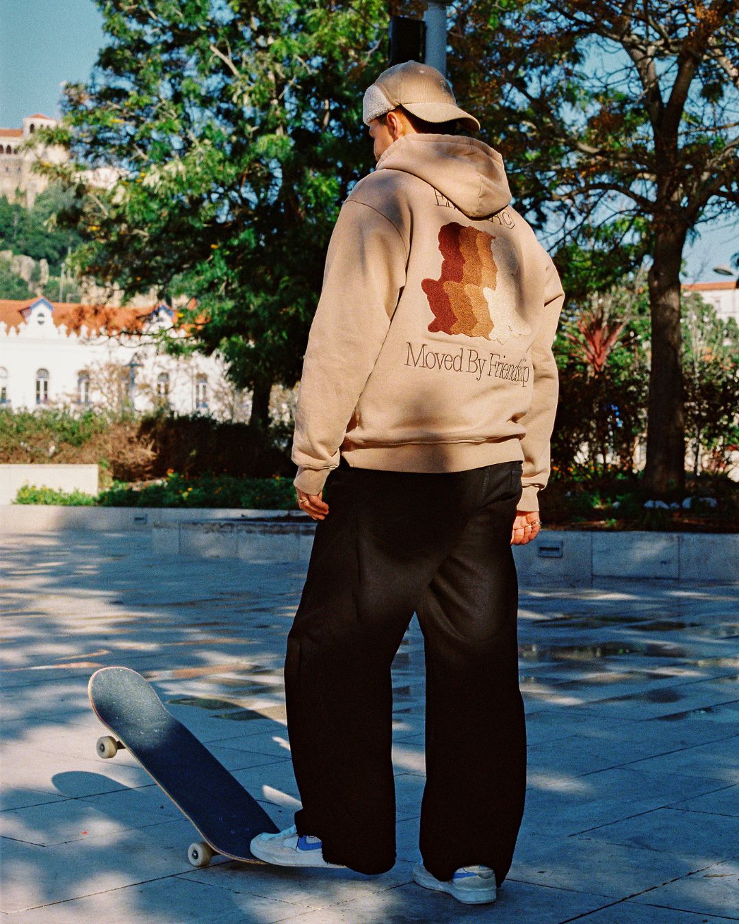 The Friendship Hoodie in camel features the motto "Moved By Friendship" embroidered on the front in a classic handwritten style. On the back, the Ikon logo appears with a dragged effect, adding a distinctive detail.