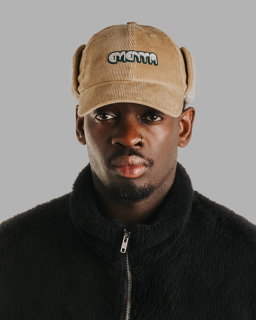 A six-panel cap constructed with a corduroy outer shell. The design includes a continuous flap extending from ear to ear with a sherpa interior for insulation and comfort, covering the back of the neck for full cold-weather protection. The flaps can be worn up or down, allowing for adjustable coverage. The cap features a curved brim for structure and coverage. The front panel displays the Dots Ementa logo.A six-panel cap constructed with a corduroy outer shell. The design includes a continuous flap extendin