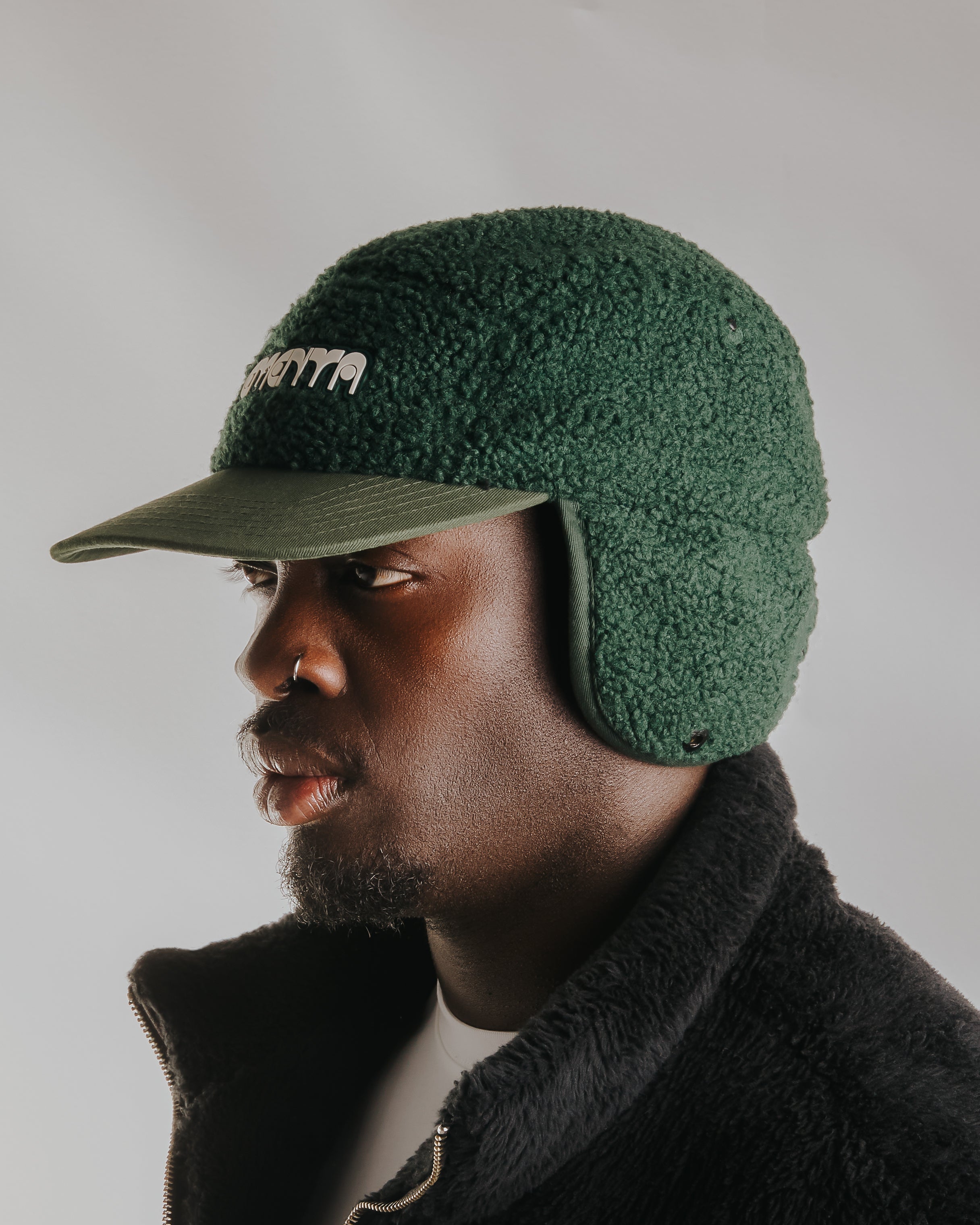 The Oreo Cap is a six-panel cap constructed with a sherpa outer shell for insulation and texture. It features ear flaps with a polar fleece lining on the inside for additional warmth and comfort. The flaps can be worn up or down, allowing for adjustable coverage. The cap has a curved brim that maintains its shape. The front panel displays the Dots Ementa logo.